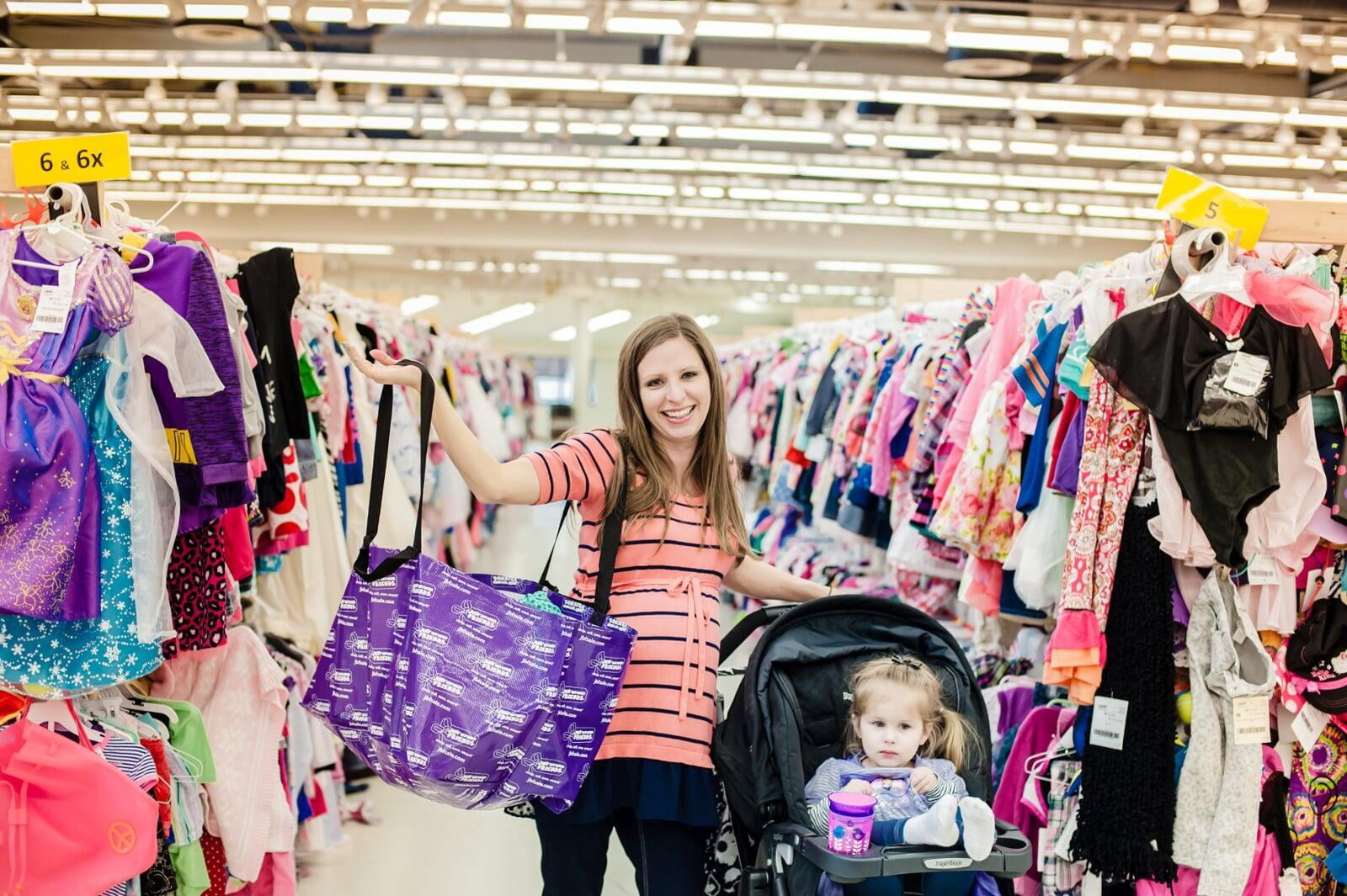 A mom with a large JBF shopping bag on her shoulder stands beside her husband who wears their toddler at a JBF sale.